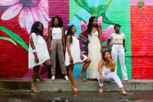 Black women of various shades and shapes posing against a colorful brick wall.