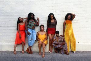 Black women of various shades and shapes posing against a white brick wall.